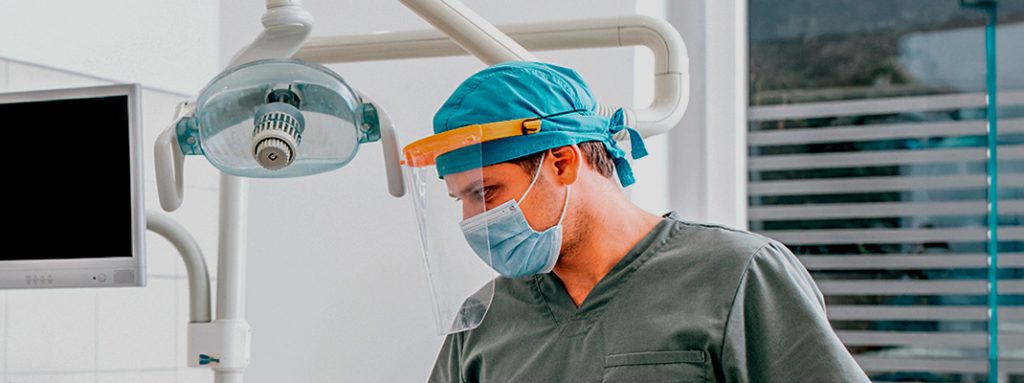 Um dentista de uniforme e máscara, com viseira. Julho Neon: A Relação entre Saúde Bucal e Saúde Geral.