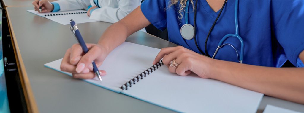 Uma mulher de uniforme azul escreve em um caderno espiral. Ele tem um estetoscópio ao pescoço. Polo Educacional EAD: O que é, Como Funciona e Qual Sua Importância.