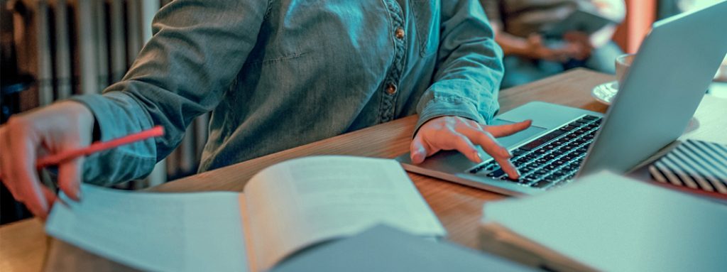 Uma pessoa escreve em um caderno e usa o laptop em uma mesa com xícara ao lado. Curso: Como Escolher a Pós-graduação Ideal: 6 Dicas para Acertar na Decisão.