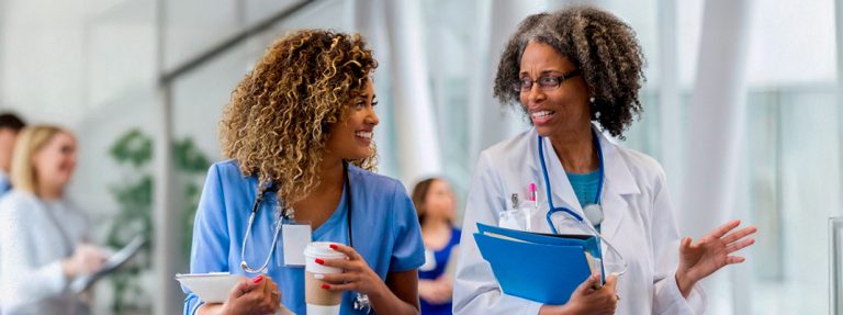 Duas profissionais da saúde conversam em um hospital. Qual é o Papel do Médico Infectologista e do Enfermeiro na Busca da Melhoria da Prática Clínica.