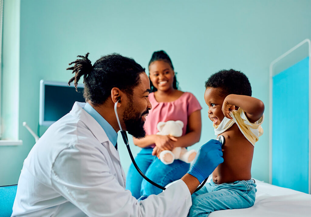 Médico da atenção primária à saúde (aps) usa o estetoscópio para fazer a auscultação cardíaca de uma criança. A mãe está sorridente ao fundo.
