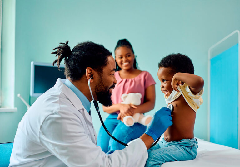 Médico da atenção primária à saúde (aps) usa o estetoscópio para fazer a auscultação cardíaca de uma criança. A mãe está sorridente ao fundo.