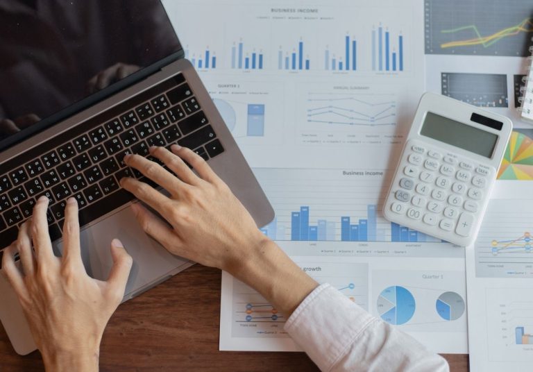 Pessoa usa laptop e calculadora sobre gráficos financeiros em mesa de madeira. Curso: Gestão de Sinistralidade.