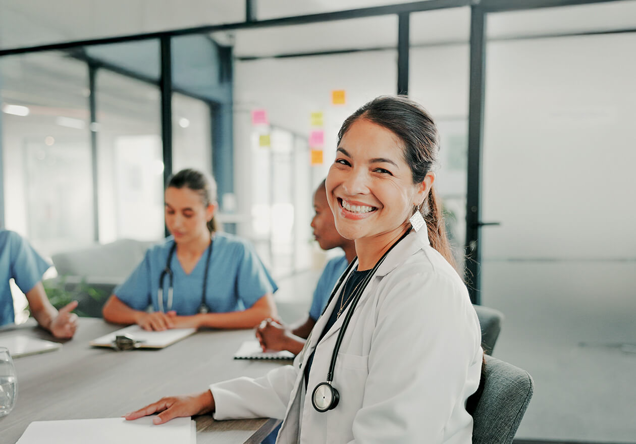 Médica sorridente em reunião com equipe sobre gestão hospitalar ead, com estetoscópio e colegas ao fundo em uma sala bem iluminada.