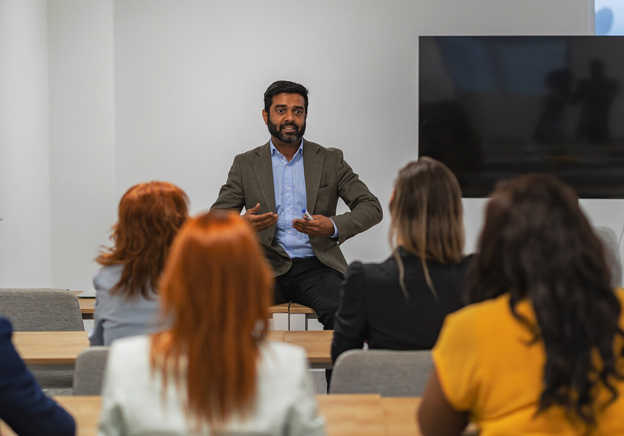 Profissional da gestão hospitalar presencial falando para público em sala de aula, com tela ao fundo, em um ambiente de treinamento ou workshop.