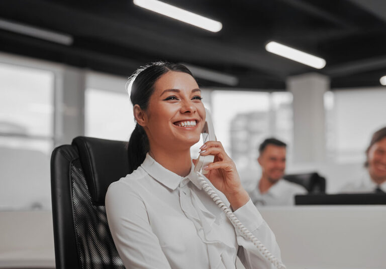 Mulher sorridente, formada no curso de secretariado, atendendo telefone em escritório, com colegas ao fundo em um ambiente bem iluminado.