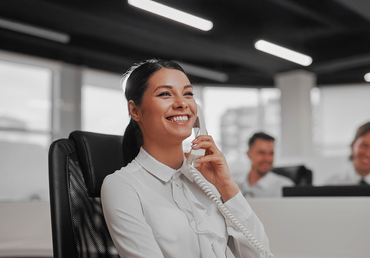 Mulher sorridente, formada no curso de secretariado, atendendo telefone em escritório, com colegas ao fundo em um ambiente bem iluminado.