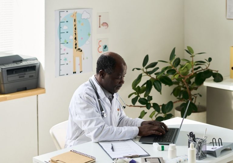 Um médico de jaleco trabalha em um laptop em seu consultório com planta e impressora ao fundo. Curso: A Dinâmica da Saúde Suplementar e o Papel do Médico Cooperado.