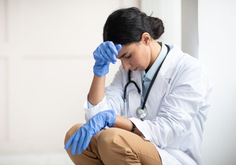 Uma médica de jaleco, com luvas azuis, está sentada e pensativa, com estetoscópio no pescoço. Curso: Abordagem à 2ª Vítima de Evento Adverso.