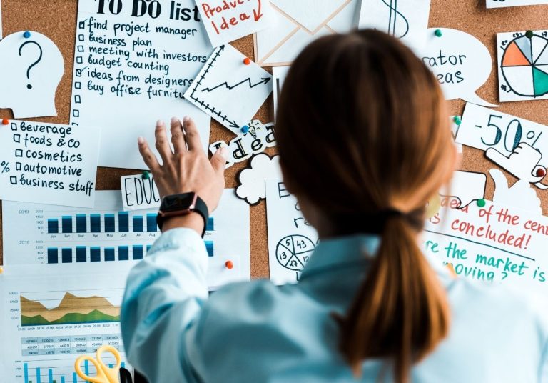 Uma mulher analisa um quadro com gráficos, listas e anotações coloridas. Curso: Analisar Problemas e Tomar Decisões.