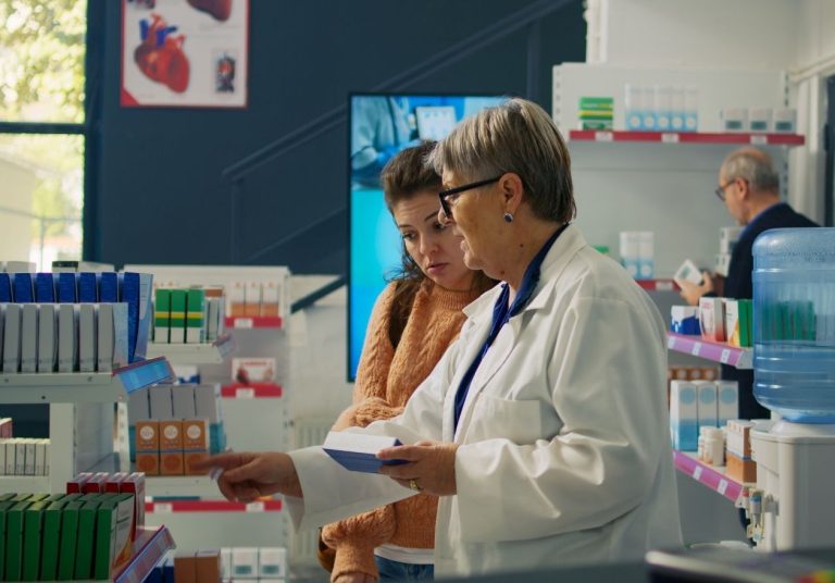 Uma farmacêutica de jaleco entrega um documento a uma cliente em uma farmácia com prateleiras ao fundo. Curso: Aperfeiçoamento em Farmácia.