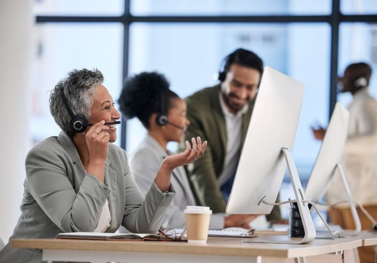 Pessoas em escritório com fones de ouvido atendem chamadas e trabalham em computadores. Curso: Atender Telefone e Responder E-mails Profissionalmente.