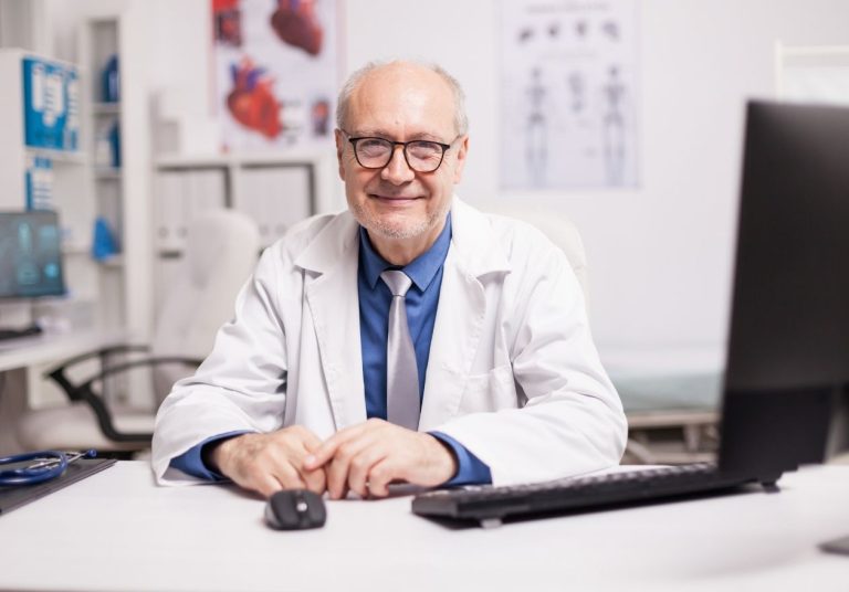 Médico em consultório com jaleco, sentado à mesa com computador e cartazes anatômicos ao fundo. Curso: Atualização Médica Personalizada.