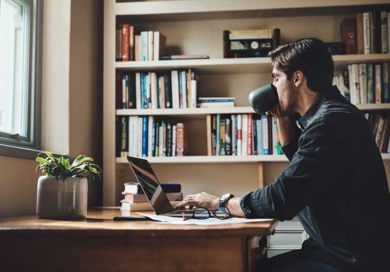 Homem trabalha em laptop e toma café. No fundo, uma estante com vários livros. Curso: Balancear a Vida.