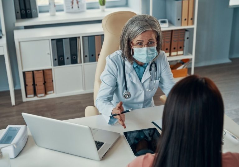 Médica com máscara conversa com paciente em consultório, com laptop e tablet na mesa. Curso: Bootcamp: Faça os Pacientes Dizerem Sim.
