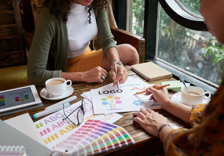 Pessoas trabalham em mesa com papéis coloridos, tablet e café. Curso: Branding.