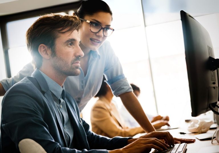Homem e mulher trabalham em computador no escritório, um orienta o outro. Curso: Código de Conduta Unimed.