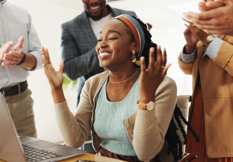 Pessoas celebram em escritório com laptop, aplaudindo e sorrindo. Curso: Competências Emocionais.