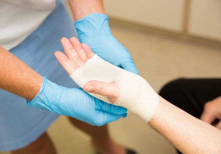 Um profissional de saúde com luvas azuis aplica uma bandagem na mão de um paciente. Curso: Condutas de Enfermagem no Tratamento de Feridas.