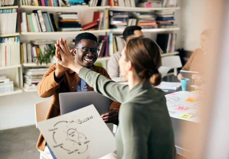 Duas pessoas sorridentes batem as mãos em um escritório com estantes de livros ao fundo. Curso: Construir Confiança.