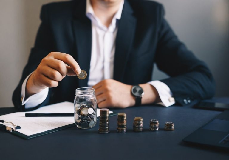 Um homem de terno coloca uma moeda em um pote de vidro com outras moedas enfileiradas na mesa. Curso: Construir Patrimônio com Qualquer Salário.
