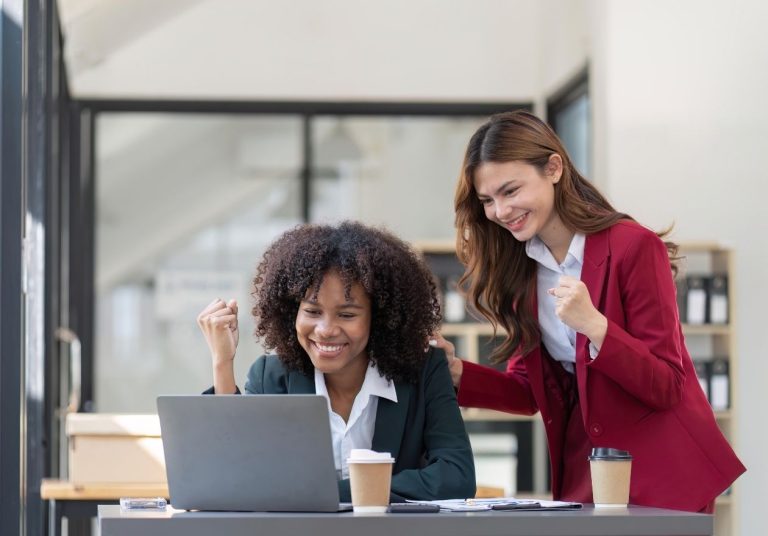 Duas mulheres celebram em um escritório, uma sentada ao laptop e outra de pé. Curso: Construir Significado no Trabalho.