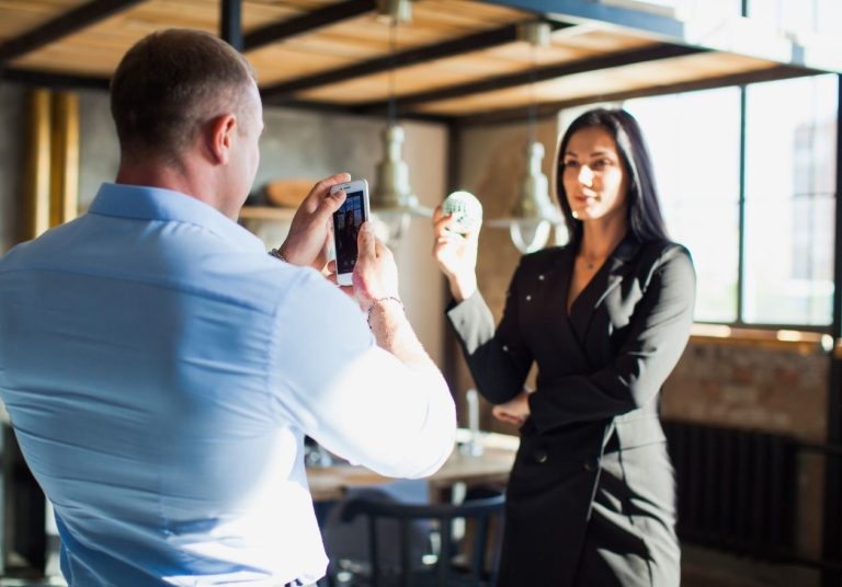 Um homem fotografa com um celular uma mulher posando com um produto em um ambiente moderno. Curso: Construir um Marketing Pessoal.