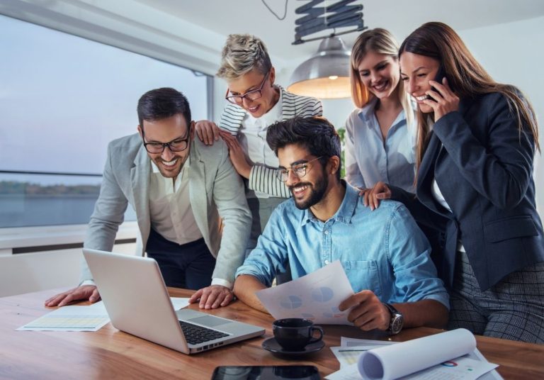 Um grupo de pessoas colabora em um escritório, com laptop, papéis e uma vista ampla pela janela. Curso: Construir uma Cultura de Engajamento.