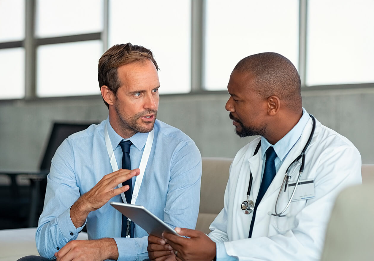 Médico segurando tablet conversa com profissional de consultoria e avaliação em serviços de saúde em um ambiente de consultório.