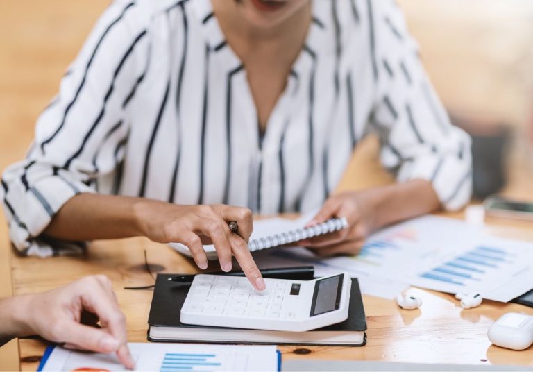 Uma mulher analisa gráficos e papéis financeiros em uma mesa com calculadora e caderno. Curso: Contabilidade para Não Contadores.