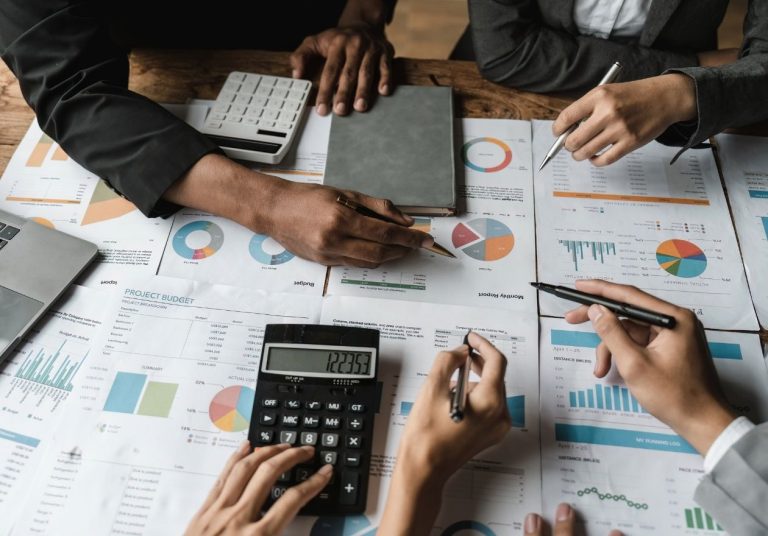 Mãos de várias pessoas analisam gráficos financeiros e usam calculadora sobre uma mesa. Curso: Contabilidade para Operadoras de Planos de Saúde.