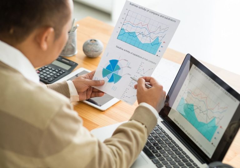 Um homem analisa gráficos financeiros em papéis e no laptop em uma mesa com calculadora. Curso: Controladoria.