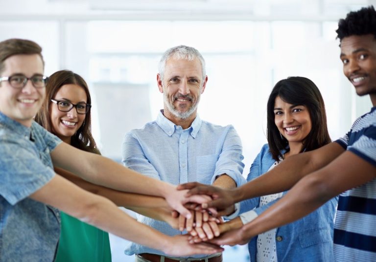 Um grupo de cinco pessoas une as mãos em círculo, simbolizando união, em um ambiente bem iluminado. Curso: Cooperativismo e Sistema Unimed.