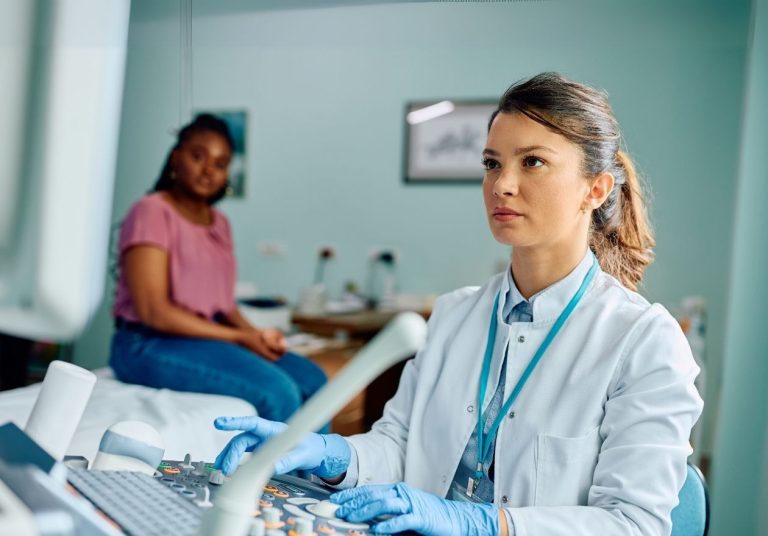Uma médica de jaleco com luvas azuis usa um equipamento em um consultório, com paciente ao fundo. Curso: Cultura de Segurança do Paciente.