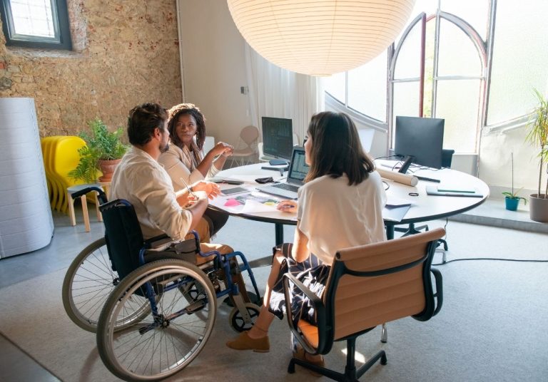 Três pessoas, uma em cadeira de rodas, conversam em uma sala com mesa redonda e laptops. Curso: Cultura e Pessoas.