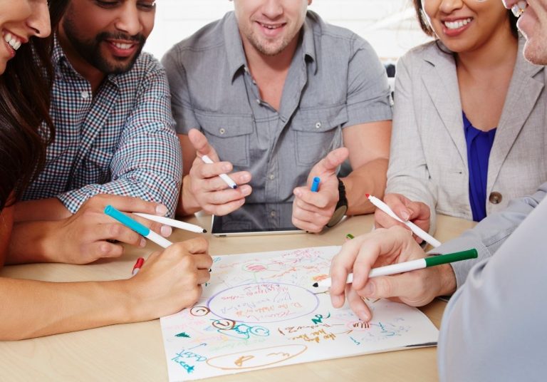 Pessoas colaborando em um desenho colorido com marcadores, estão debruçados em uma mesa. Curso: Desenvolver a Sua Criatividade.