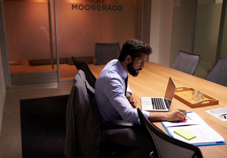 Homem trabalha em um laptop em uma sala de reunião, com papéis e caneta na mesa. Curso: Desenvolver Disciplina para Alcançar Metas.