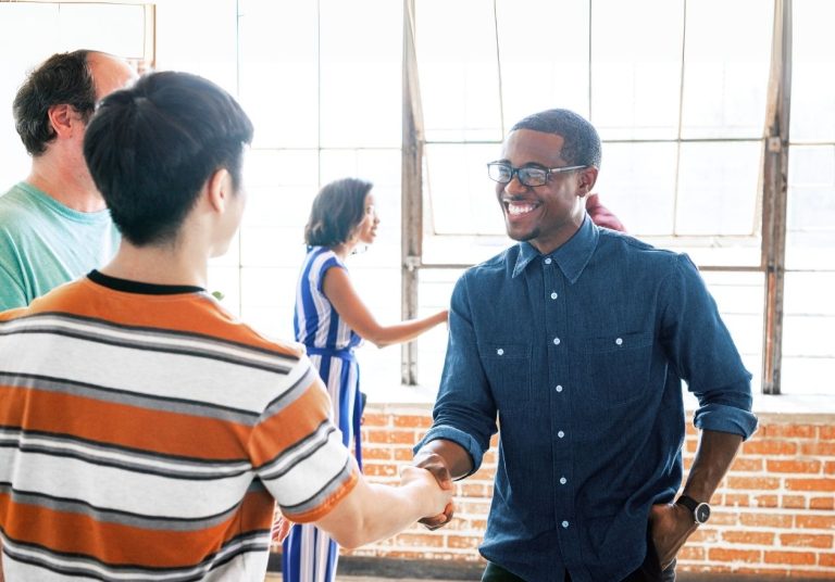 Duas pessoas se cumprimentam com um aperto de mão em um ambiente de escritório, enquanto outras conversam ao fundo. Curso: Desenvolver Inteligência Social.