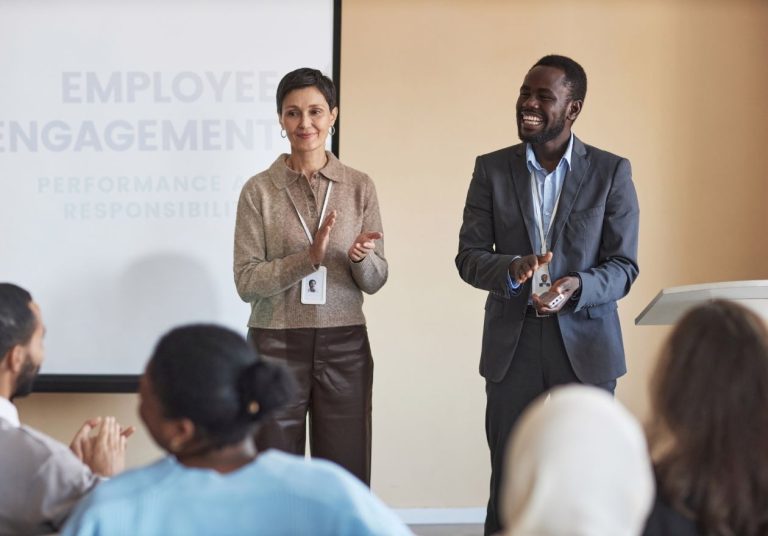 Dois palestrantes falam para uma audiência em uma sala com uma tela ao fundo exibindo. Curso: Desenvolvimento de Líderes com Foco na Qualidade.