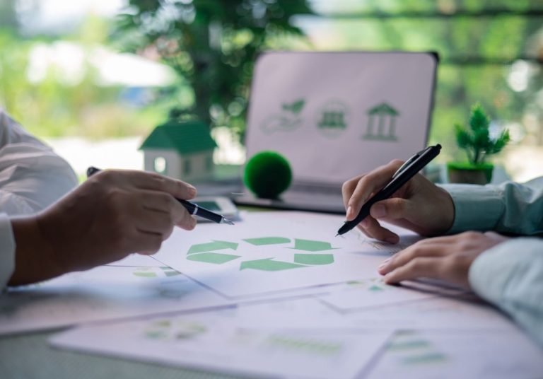 Duas pessoas trabalham em um projeto com um símbolo de reciclagem, em uma mesa com laptop e plantas. Curso: Desenvolvimento Sustentável.