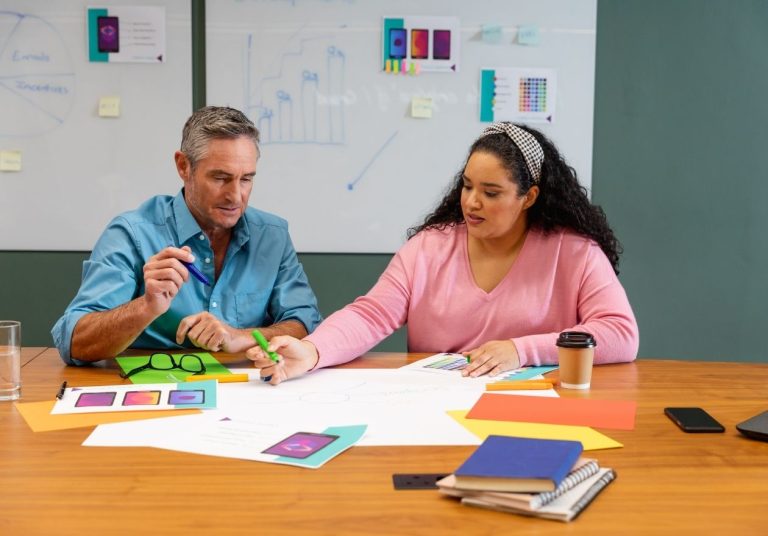 Duas pessoas discutem ideias em uma mesa com papéis coloridos, canetas e quadro branco ao fundo. Curso: Design Estratégico.