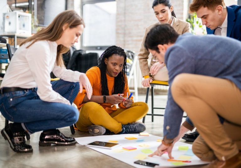 Grupo de pessoas sentadas no chão, colaborando com papéis e tablets em um ambiente descontraído. Curso: Design Thinking nos Negócios.