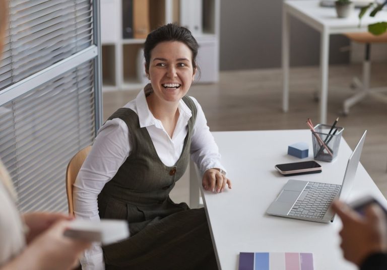 Mulher sorridente sentada à mesa com laptop, em um ambiente de escritório interagindo com outras pessoas. Curso: Despertar o Entusiasmo.