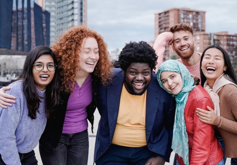 Grupo diverso de pessoas sorrindo e se abraçando ao ar livre, com prédios ao fundo. Curso: Diversidade Cultural.
