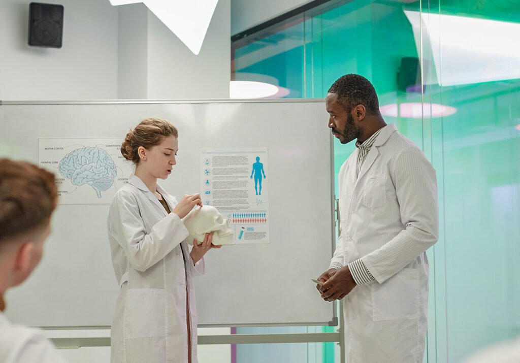 Dois médicos de jaleco conversam em frente a um quadro com diagramas do corpo humano. Curso: Docência em Saúde.
