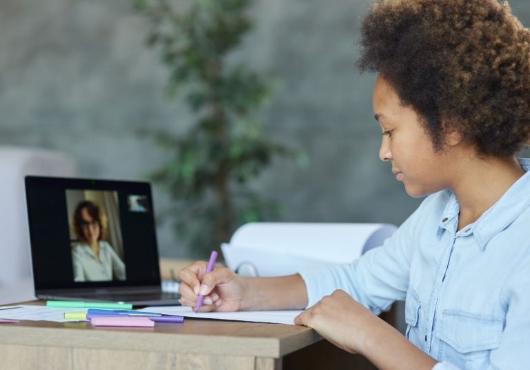 Mulher faz anotações enquanto participa de uma videoaula no laptop, em um ambiente com plantas ao fundo. Curso: Educação Cooperativista.