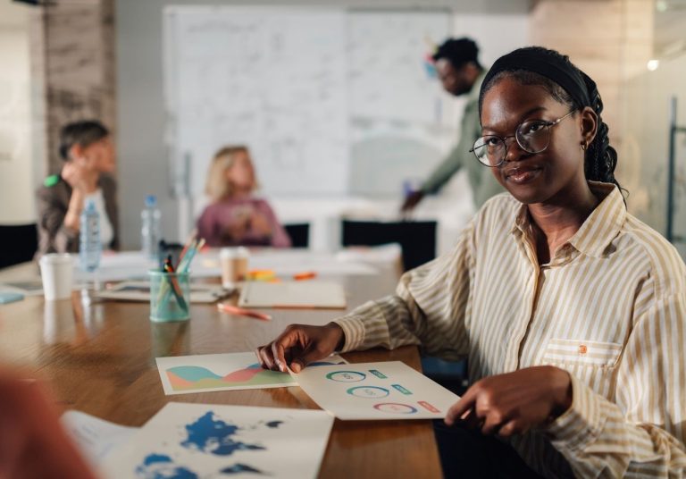 Uma mulher analisa papéis em uma mesa, com colegas ao fundo em um ambiente de trabalho. Curso: Empowerment.