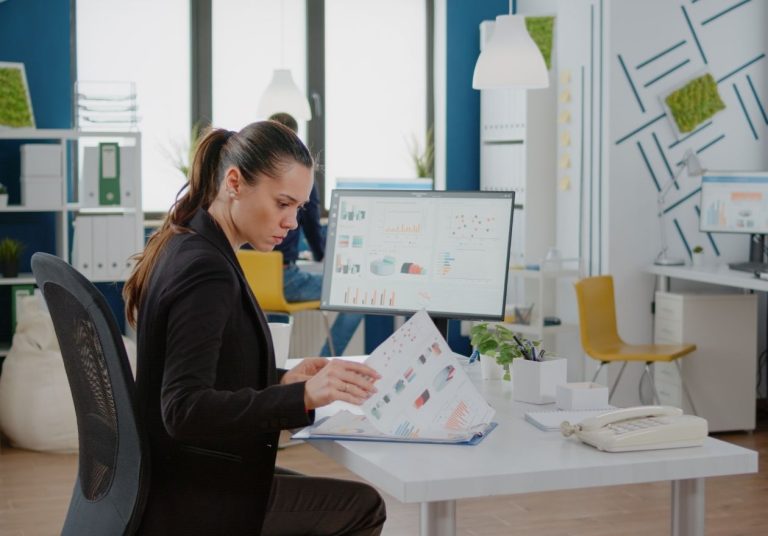 Uma mulher trabalha em um computador com papéis e gráficos em um escritório moderno. Curso: Empreendedorismo.