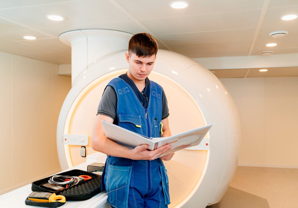Homem, especialista em engenharia clínica, está revisando manual ao lado de uma máquina de ressonância magnética em ambiente hospitalar, com ferramentas sobre a mesa.