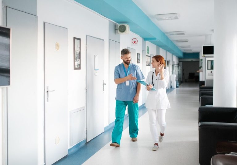 Dois profissionais de saúde conversam em um corredor de hospital com portas ao lado. Curso: Entendendo o SBA e a Metodologia ONA.